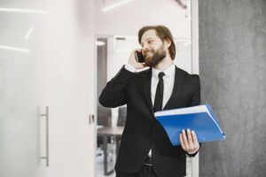 Smiling businessman in suit at office holding documents and using smartphone.